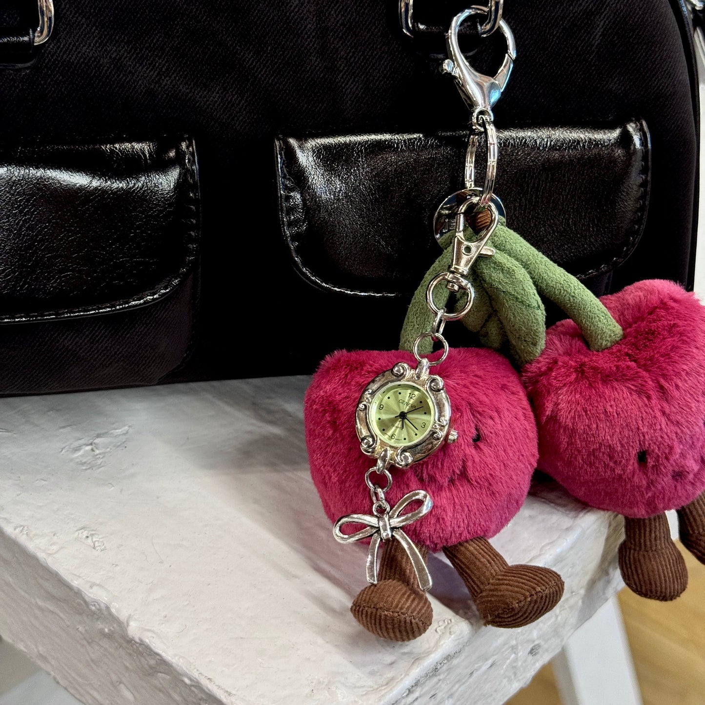 A close-up of a black handbag with a novelty keychain charm shaped like two pink cherries, each with a small round watch and green leaves, resting on a white wooden surface.